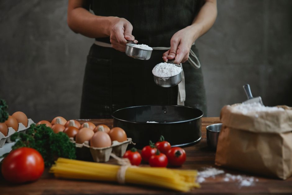 Crop faceless person in black apron standing near counter and adding flour in bowl while making homemade pasta with eggs and spaghetti near tomatoes and herbs