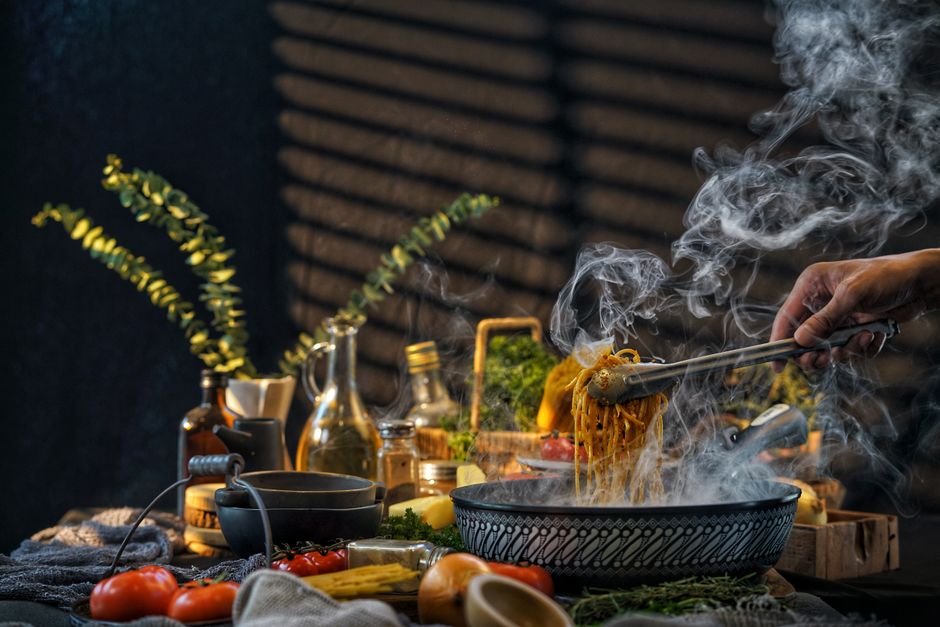 A steaming bowl of pasta being served in a well-lit rustic kitchen setting with fresh ingredients and utensils.
