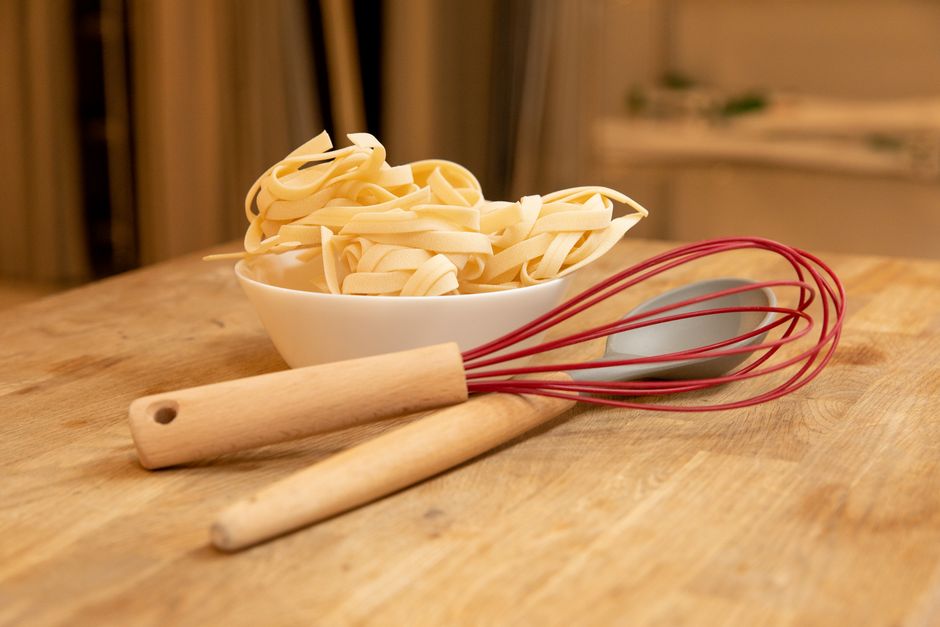 Wooden kitchen table with uncooked pasta in a bowl and utensils, ideal for cooking themed visuals.